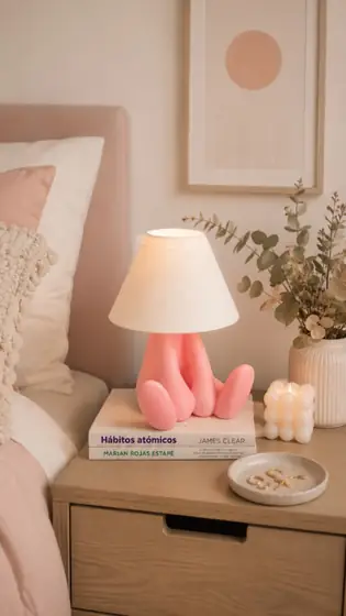Lámpara con base rosa en forma de figura sentada y pantalla clara sobre una mesa de noche