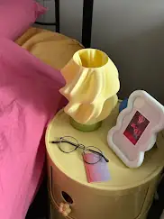 Lámpara amarilla con pantalla ondulada y base verde sobre una mesa junto a una cama