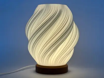 Lámpara encendida con luz cálida, pantalla blanca en espiral y base café, frente a un fondo azul